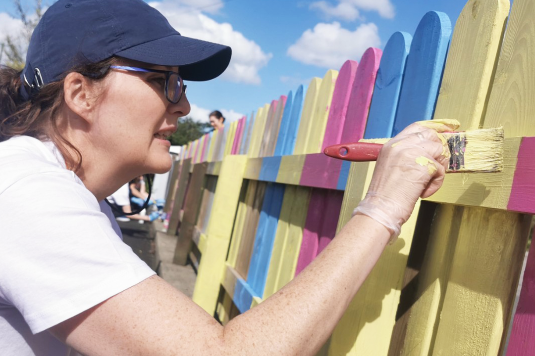 A woman wearing a peek cap and t-shirt painting a multicolored wooden fence to help a community